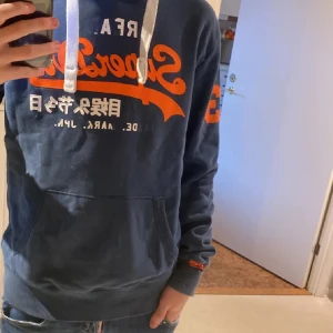 Blå hoodie från Superdry - Säljer en snygg blå hoodie från Superdry med orange och vit text på bröstet. Den har en justerbar huva med vita snören och en stor ficka framtill. Perfekt för en avslappnad stil. 🚨PRIS KAN DISKUTERAS🚨