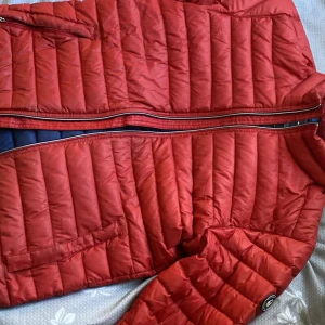 Röd dunjacka från Sail Racing - Snygg röd dunjacka från Sail Racing med dragkedja och quiltad design. Jackan har en kontrasterande blå insida och praktiska fickor med dragkedjor. Perfekt för kyliga dagar när du vill hålla stilen. 🧥