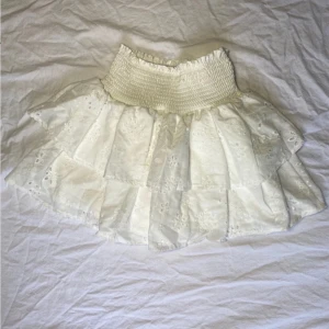 Vit kjol med volanger från Shein - Charmig vit kjol från Shein med flera lager av volanger och ett elastiskt midjeband. Perfekt för en somrig look. Kjolen har ett vackert broderat mönster som ger en extra touch av elegans.