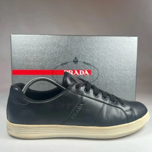 Svarta sneakers från Prada - Snygga svarta sneakers från Prada i skinn med klassisk snörning. Skorna har en rund tå och en vit sula som ger en stilren kontrast. Perfekta för en stilmedveten look.