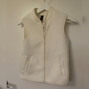 Vit fleeceväst från SOC - Säljer en vit fleeceväst från SOC med dragkedja framtill. Västen har en enkel och stilren design med två fickor. Västen är i teddy-material, storlek 134/140. Mycket bra skick. 