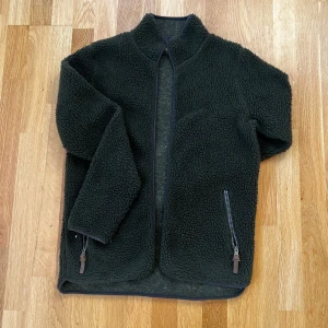 Grön teddyjacka från Strovå - Mysig grön teddyjacka från Strovå. Perfekt för kyligare dagar med sin fluffiga och varma design. Jackan har en hög krage och långa ärmar för extra värme. 