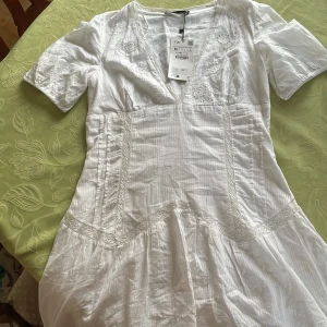 Vit klänning med broderi - Säljer en vacker vit klänning med broderade detaljer och v-ringning. Klänningen har korta ärmar och en feminin passform. 