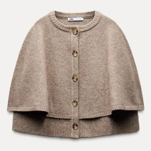 Beige cape från Zara - Säljer en stilren beige cape från Zara med knappar framtill. Den har en rund hals och ribbade kanter som ger en elegant touch. Perfekt för lager-på-lager under kyliga dagar. Storlek M. Aldrig 2 ggr. 