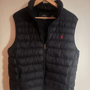 Mörkblå dunväst från Polo Ralph Lauren - Snygg mörkblå dunväst från Polo Ralph Lauren med dragkedja och broderad logga i rött på bröstet. 