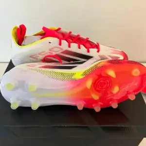 Snygga Adidas F50 fotbollsskor i vitt med svarta och röda detaljer. Skorna har en modern design med snörning och en färgglad sula i rött och gult. Perfekta för att dominera på planen med stil.