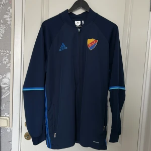 Mörkblå träningsjacka från Adidas - Säljer en mörkblå träningsjacka från Adidas med DIF-logga på bröstet. Jackan har långa ärmar och dragkedja framtill. Den har ljusblå detaljer längs ärmarna och sidorna. 