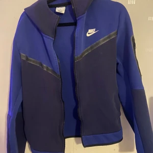 Blå jacka från Nike - Säljer en blå jacka från Nike med dragkedja och vit logga på bröstet. Jackan har långa ärmar och en huva. Perfekt för sportiga aktiviteter eller som en casual vardagsjacka.