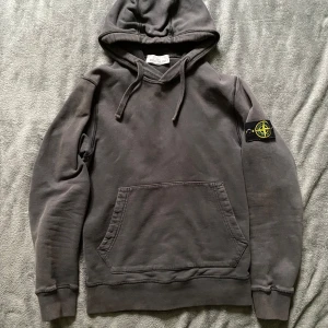 Hoodie från Stone Island - Säljer en snygg hoodie från Stone Island med klassisk logga på ärmen. Tröjan har en stor ficka framtill och justerbar huva med dragsko. Perfekt för en avslappnad stil. Den är välanvänd, men har tappat lite av sin färg som man ser på bilderna. Köpt här på Plick så har inget bevis på att den är äkta, men tror att den är det. Vid frågor eller funderingar skicka ett dm💫