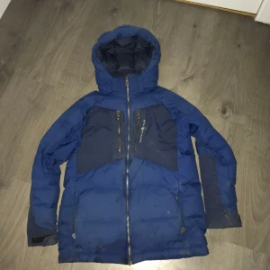 Blå dunjacka från Sail Racing - Säljer en blå dunjacka från Sail Racing med huva och dragkedja. Jackan har två bröstfickor med dragkedjor och är tillverkad i nylon. Perfekt för kalla dagar. Jackan behövs tvättas