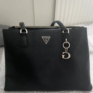 Svart axelväska från Guess - Snygg svart axelväska från Guess med elegant design. Insidan är rymlig med flera fack för enkel organisering. Använd 2-3 gånger. Säljer pga att jag köpte en annan väska. 