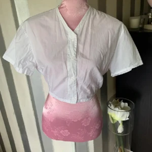 Vit kortärmad blus - Säljer en vit kortärmad blus med knappar framtill. Blusen är croppad och har en enkel design som passar perfekt för varma dagar. Kort i modellen 