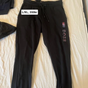 Svarta mjukisbyxor från Hugo Boss - Svarta mjukisbyxor från Boss med elastisk midja och snörning. Byxorna har en logotyp på sidan och är perfekta för en avslappnad stil. De är tillverkade i ett mjukt material för extra komfort. Collab med NBA