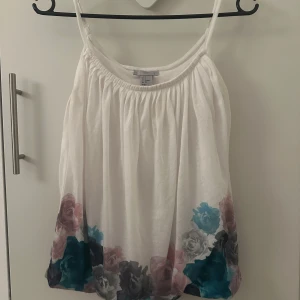 Vit blommig linnetopp - Säljer en söt vit linnetopp med tunna axelband och ett blommigt mönster i rosa, blått och grått längs nederkanten. Perfekt för sommardagar! 🌸