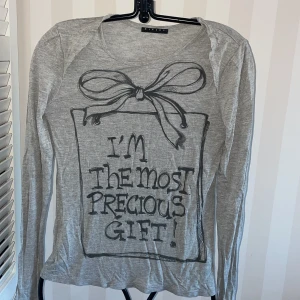 Grå långärmad tröja från Sisley st.170/XS - Säljer en grå långärmad tröja från Sisley med ett tryck framtill som säger 'I'm the most precious gift!' och en stor rosett. Perfekt för en avslappnad stil.