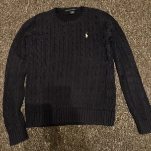 Mörkblå stickad tröja från Ralph Lauren äkta - Säljer en stilren mörkblå stickad tröja från Ralph Lauren med kabelstickat mönster. Tröjan har långa ärmar och en liten broderad logotyp på bröstet. Perfekt för kyligare dagar.