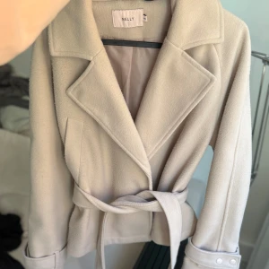 Beige kappa - Beige kort kappa från Nelly med klassisk krage och knytskärp i midjan. Jackan har långa ärmar med dekorativa knappar vid ärmsluten. 