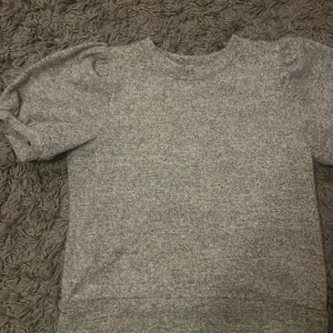 Grå kortärmad tröja - Säljer en stilren grå tröja med korta ärmar och puffiga axlar. Perfekt för en avslappnad look. Tröjan har en rund halsringning och är tillverkad i ett mjukt material. Använd 2-3 gånger.