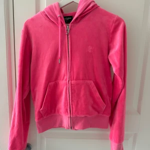 Rosa hoodie från Juicy Couture - Säljer en snygg rosa hoodie från Juicy Couture med dragkedja och broderad logga på bröstet. Den har en mysig känsla och kängurufickor framtill. Perfekt för en avslappnad stil.