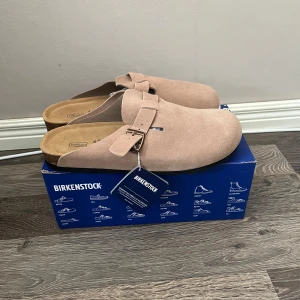 Beige Birkenstock Boston tofflor i mocka - Säljer ett par beige Birkenstock Boston tofflor i mjuk mocka med justerbart spänne. Skorna har en klassisk rund tå och svart mönstrad sula. Perfekta för en avslappnad och stilren look.