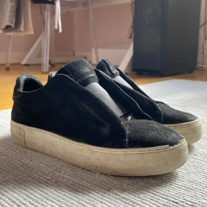 Svarta sneakers från Axel Arigato - Svarta sneakers från Axel Arigato med minimalistisk design och vit platt sula. Skorna är i mocka och har en rund tå utan synlig snörning för en clean look. Perfekta för dig som gillar stilrena och enkla skor. Jag har box och en liten vätska till skorna som man lägger in skorna i !