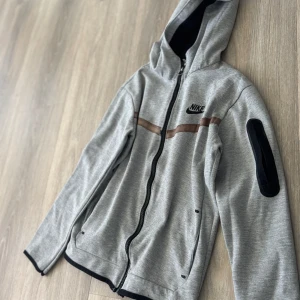 Grå zip-hoodie från Nike - Grå hoodiejacka från Nike med svart dragkedja, huva och svarta detaljer. Jackan har en brun rand över bröstet och en svart ficka på ena ärmen. Perfekt för en sportig stil.