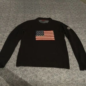 Blå stickad tröja med amerikansk flagga från Gant - Mörkblå stickad tröja från Gant med amerikansk flagga broderad på bröstet och texten 'GANT NEW HAVEN 1949' på ena ärmen. Klassisk rund halsringning och ribbade muddar vid ärmslut och nederkant. Den har en slitning på höger arm men annars är den i bra skick!