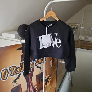 Svart croppad sweatshirt från Love Moschino - Säljer en svart croppad sweatshirt från Love Moschino med stort vitt tryck framtill där det står 'Love'. Tröjan har rund halsringning och långa ärmar. Perfekt statement-plagg för dig som gillar streetwear.