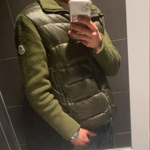 Grön Cardigan från Moncler med stickade ärmar - Snygg grön dunväst från Moncler med stickade ärmar och hög krage. Västen har dragkedja framtill med röd, vit och blå detalj samt Moncler-logga på ärmen. Perfekt för lager-på-lager och stilren look.