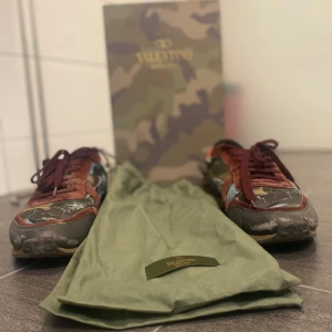 Valentino Rockrunner sneakers med camouflage - Säljer ett par Valentino Rockrunner sneakers med camouflage-mönster i grönt, brunt och svart. Skorna har detaljer i röd mocka, svarta och blå inslag samt klassisk snörning. Yttersulan är beige och har Valentino-logga undertill. Kommer med originalpåse, påse, kvitto och extra snören