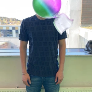 Mörkblå fendi t-shirt  - Säljer en mörkblå fendi t-shirt med ett diskret rutmönster i tyget. T-shirten har normal passform personen på bilden är 179 68kg.