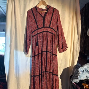 Rosa/röd  långklänning med blommigt mönster - Säljer en röd långklänning med småblommigt mönster och svarta detaljer. Klänningen har långa ärmar, v-ringning med knytband och markerade sömmar. Perfekt för en sommar kväll :D väldigt skön. Jag är M och den passar men den är en L