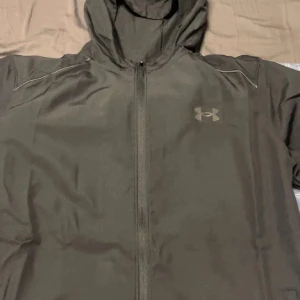 Svart träningsjacka med huva från Under Armour - Säljer en svart träningsjacka från Under Armour med huva och dragkedja framtill. Jackan har logga på bröstet och stor text på ryggen. Perfekt för träning och sportiga aktiviteter.