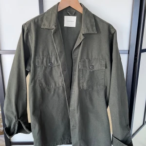 Grön overshirt från A Day's March - Säljer en stilren grön overshirt från A Day's March. Skjortan har två stora bröstfickor med knappar och klassisk krage. Perfekt att bära öppen eller stängd för en avslappnad look.