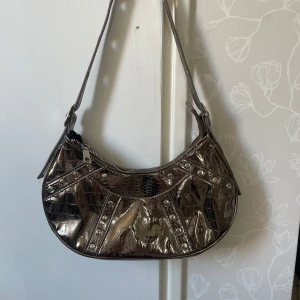 Silver axelväska  - Snygg axelväska i metallic silver med krokopräglad yta och nitar längs kanterna. Väskan har dragkedja upptill och justerbart axelband med metallringar. 
