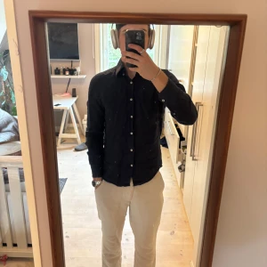 Mörkblå linneskjorta  - Säljer en stilren mörkblå linneskjorta från These Glory Days. Skjortan har slim fit-passform, klassisk krage och knäppning framtill med vita knappar. Perfekt för dig som gillar en enkel men snygg look. Nypris 1199kr. Passar främst S men M fungerar om man gillar att den sitter lite tajtare. Väldigt bra skick 10/10. Perfekt för sommarn! Skriv för frågor eller funderingar.