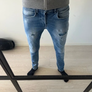 Blå slitna jeansbyxor från Dondup - Säljer ett par blå jeansbyxor från Dondup med slitningar och hål på låren och knäna. Byxorna har klassisk femficksdesign, normal passform och knappgylf. Perfekta för en avslappnad och trendig look. Tror jag har kvitto och tags kvar.