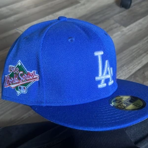 Blå LA Dodgers keps från New Era - Säljer en blå LA Dodgers 59FIFTY keps från New Era med broderad logga framtill, World Series 1988-märke på sidan och texten 'Love & Hustle 4 the Game' bak. Kepsen har platt skärm och är i bomull.