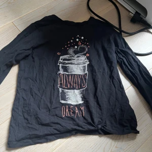 Svart långärmad tröja med tryck från ”Name It” är för barn  - Svart långärmad tröja från Name It med tryck av en kaffemugg och texten 'Always Dream' på framsidan. Tillverkad i ekologisk bomull och har en avslappnad passform. Denna tröjan är för barn mellan 9-10 år/134-140, tröjan har dock ett litet hål längst ner men de syns inte.