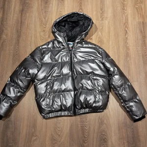 Silver pufferjacka med huva från Ombre - Snygg och glansig silverfärgad pufferjacka från Ombre med huva och dragkedja. Jackan har två stora fickor framtill och elastisk nederkant. Perfekt för kalla dagar när du vill sticka ut lite extra. Den är billigt eftersom den var använt en gång och jag vill inte ha den längre