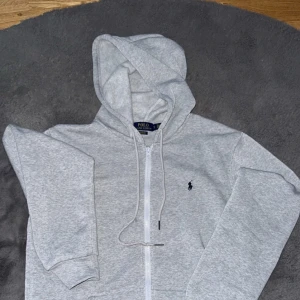 Grå hoodie från Polo Ralph Lauren - Säljer en klassisk ljusgrå hoodie från Polo Ralph Lauren med dragkedja och huva. Tröjan har en broderad logga på bröstet och två fickor framtill. Perfekt för en avslappnad stil.
