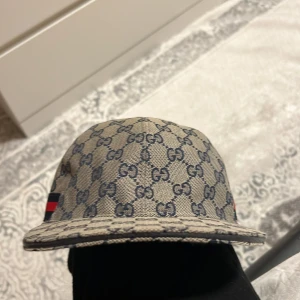 Gucci keps med monogram och röd-blå rand - Säljer en keps från Gucci med klassiskt GG-monogram i grått och blått. Kepsen har en röd-blå rand på sidan och justerbart svart läderband bak. Perfekt för dig som gillar lyxiga accessoarer med tydlig logga. Storlek M och kommer med Gucci påsen!