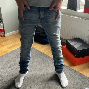 Ljusblå jeans från Nudie Jeans - Säljer ett par ljusblå jeans från Nudie Jeans med snygga slitningar och detaljerade sömmar på bakfickorna. Jeansen har klassisk femficksdesign och normal / slim passform. Perfekta för dig som gillar en stilren och avslappnad look.