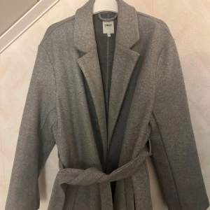 Grå kappa med bälte från ONLY - Snygg grå kappa från ONLY med klassisk krage och bälte i midjan. Långärmad modell med rak passform, perfekt för kyligare dagar. Enkel och stilren design som passar till många olika outfits. Använt Max 2 gånger 
