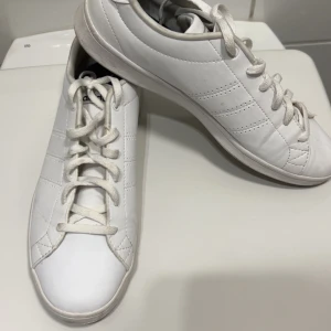 Vita sneakers från Adidas - Säljer ett par klassiska vita sneakers från Adidas med diskreta ränder på sidorna och snörning framtill. Skorna har en låg profil och är tillverkade i syntetmaterial med platt sula. Perfekta för en stilren och enkel look.