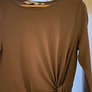 Brun långärmad klänning med knytdetalj från Kappahl - Säljer en stilren brun långärmad klänning från Kappahl med rund halsringning och snygg knytdetalj i midjan. Perfekt för dig som gillar enkel och elegant stil.