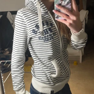 Vit och marinblå randig hoodie från Abercrombie & Fitch - Säljer en vit hoodie med marinblå ränder från Abercrombie & Fitch. Tröjan har dragkedja, huva med snören och broderad logga på bröstet och ärmen. Muddar vid ärmslut och midja. Perfekt för en avslappnad stil.