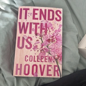 It Ends With Us - Populär roman med omslag i träliknande beige bakgrund och stora rosa bokstäver. Framsidan har ett blommotiv med rosa blommor som ser ut att spricka. Perfekt för dig som gillar samtida kärleksromaner.