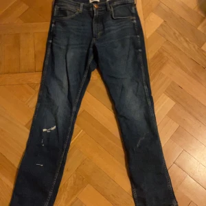Mörkblå jeans från Wrangler - Säljer ett par mörkblå jeans från Wrangler med klassisk femficksdesign och raka ben. Jeansen har snygga slitningar framtill och tydliga sömmar på bakfickorna. Perfekta för dig som gillar en tidlös denimlook.