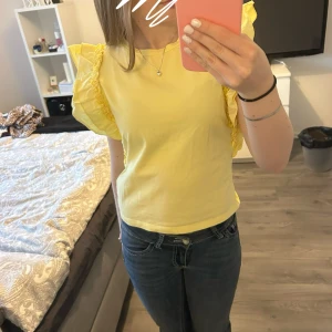 Gul topp med volangärmar - Säljer en söt gul topp med volangdetaljer på axlarna. Toppen har rund halsringning och är tillverkad i ett mjukt material som känns skönt mot huden. Perfekt för att liva upp garderoben med lite färg!